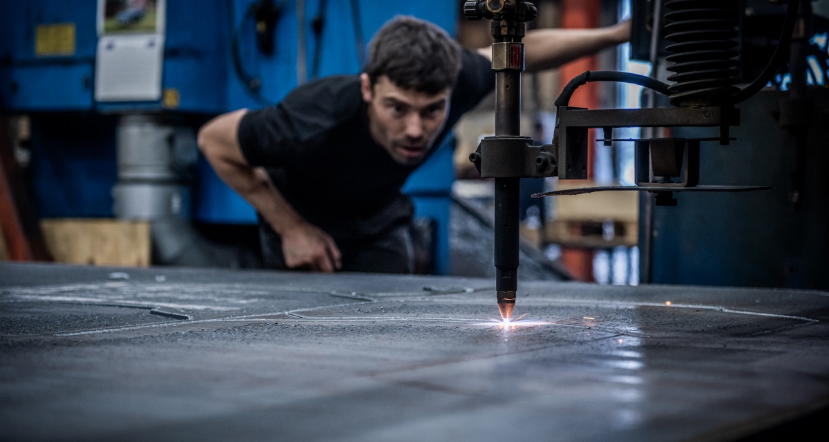 En person observerar noggrant en maskin som skär mönster till en metallplåt i en industriell miljö.