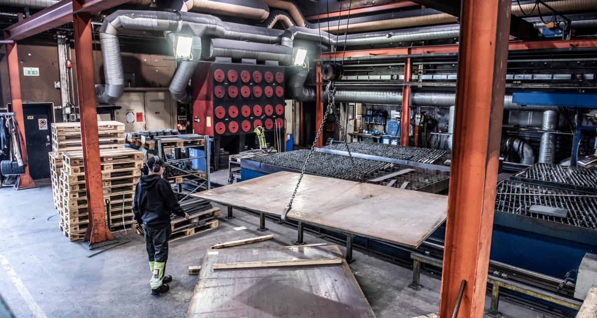 Arbetare i en industrianläggning med maskiner och utrustning, inklusive pallar och stora metallplåtar, under lysrörsbelysning.